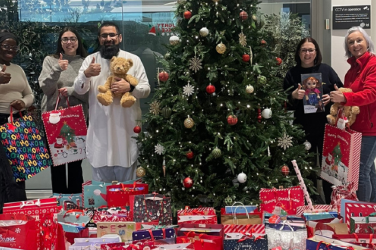 Donated presents under a tree with staff stood for a photo