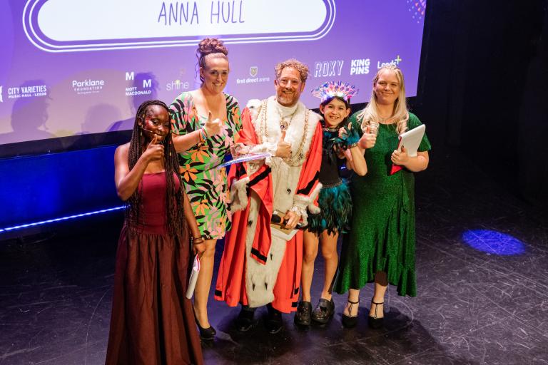 Award recipients and presenters pose on stage with a trophy beneath a screen displaying the name "ANNA HULL."