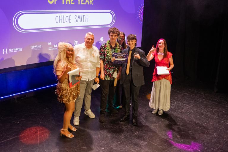 Group of award recipients and presenters on a theatre stage, with one person holding a certificate and another holding a trophy beneath a screen displaying "CHLOE SMITH."